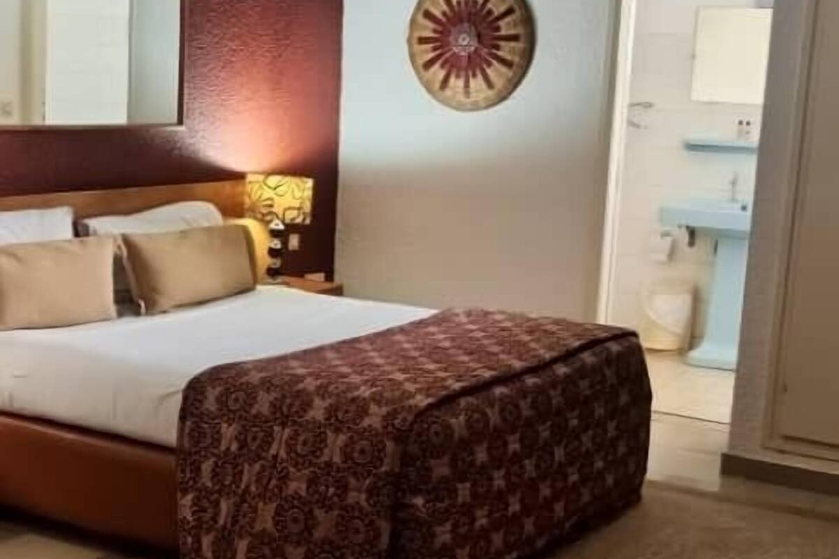 A bedroom with a bed, a bedside table, a lamp, a decorative wall piece, and a view of the bathroom.