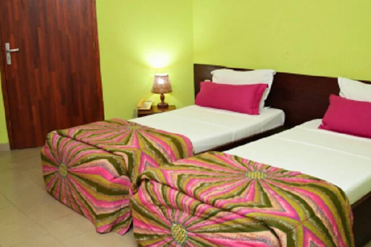 A hotel room with two beds, each with a pink and green patterned bedspread. There is a wooden door and a nightstand with a lamp.