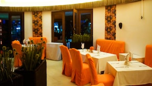 An indoor dining area with orange chairs and tables, a potted plant, and a decorative wall panel.
