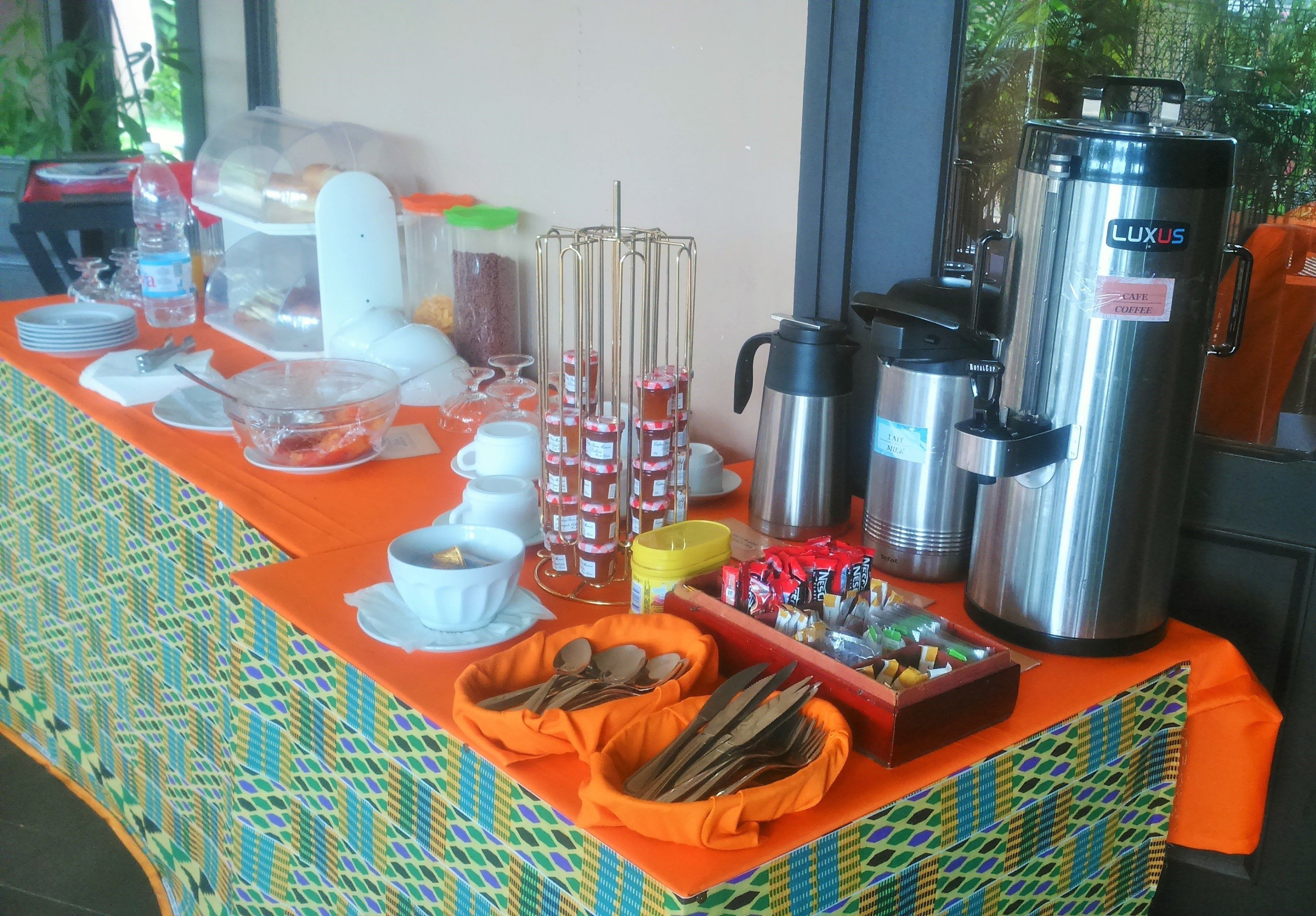 A breakfast buffet setup with various food items, utensils, and beverage dispensers.
