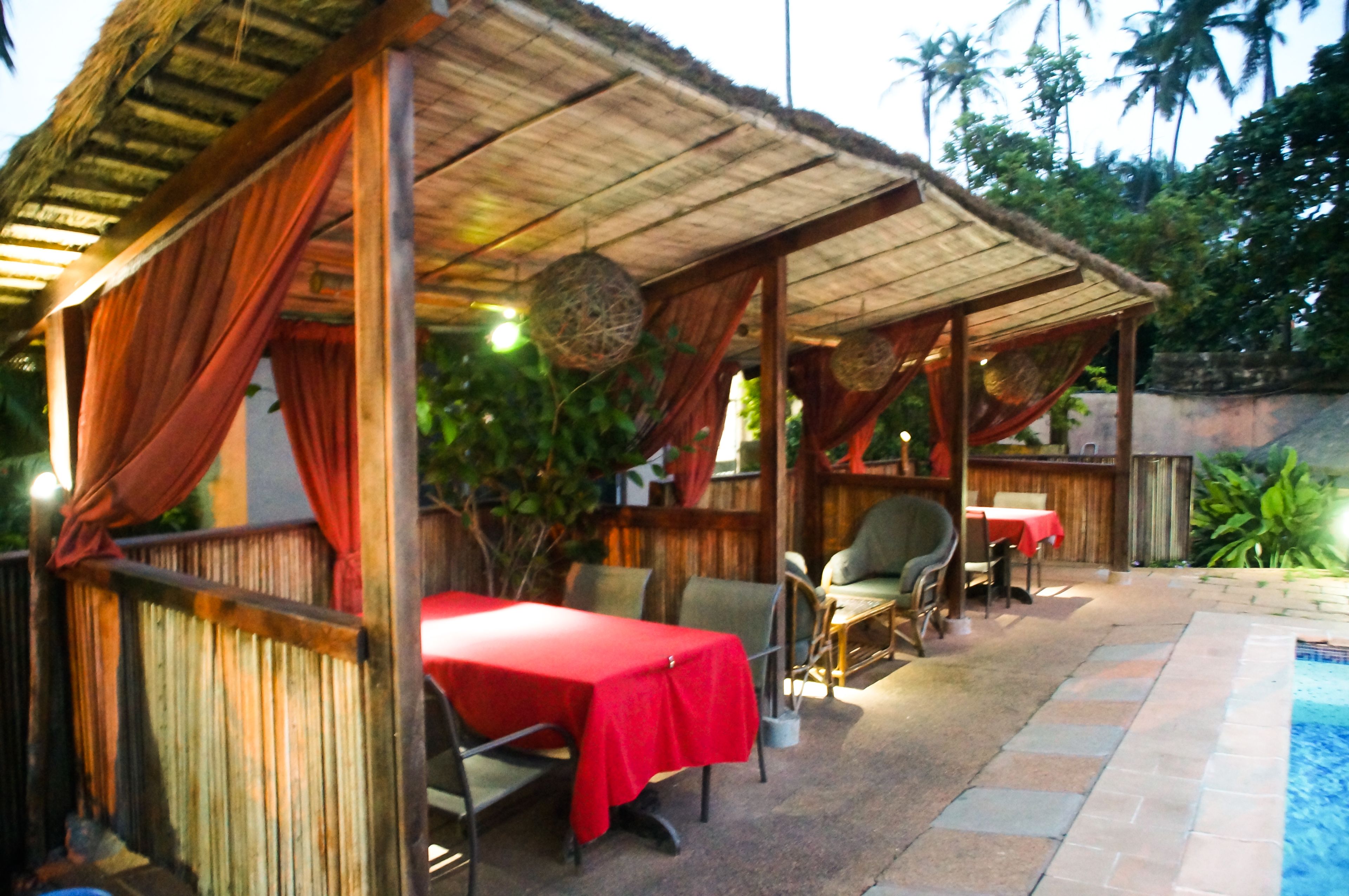 Un espace de restauration en plein air avec des tables recouvertes de nappes rouges, des chaises en bois et une structure dotée d’un toit de chaume.