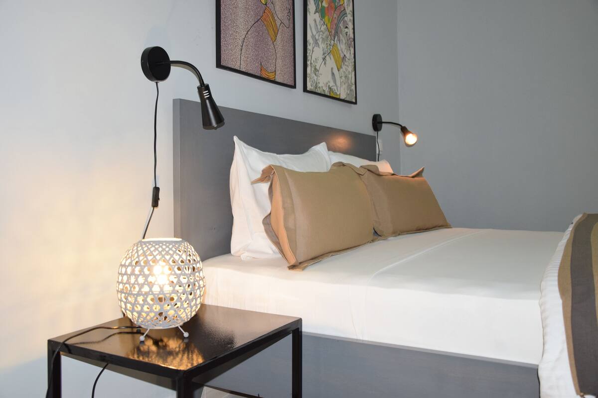 A neatly made bed with white and beige bedding, a black bedside table with a decorative lamp, and two wall-mounted lamps.