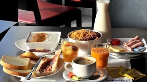 A breakfast table with bread, jam, a bowl of cereal, a plate of sausages, a glass of orange juice, a cup of coffee, and a bottle of milk. - Brit Hotel Saint-Quentin (Fayet)