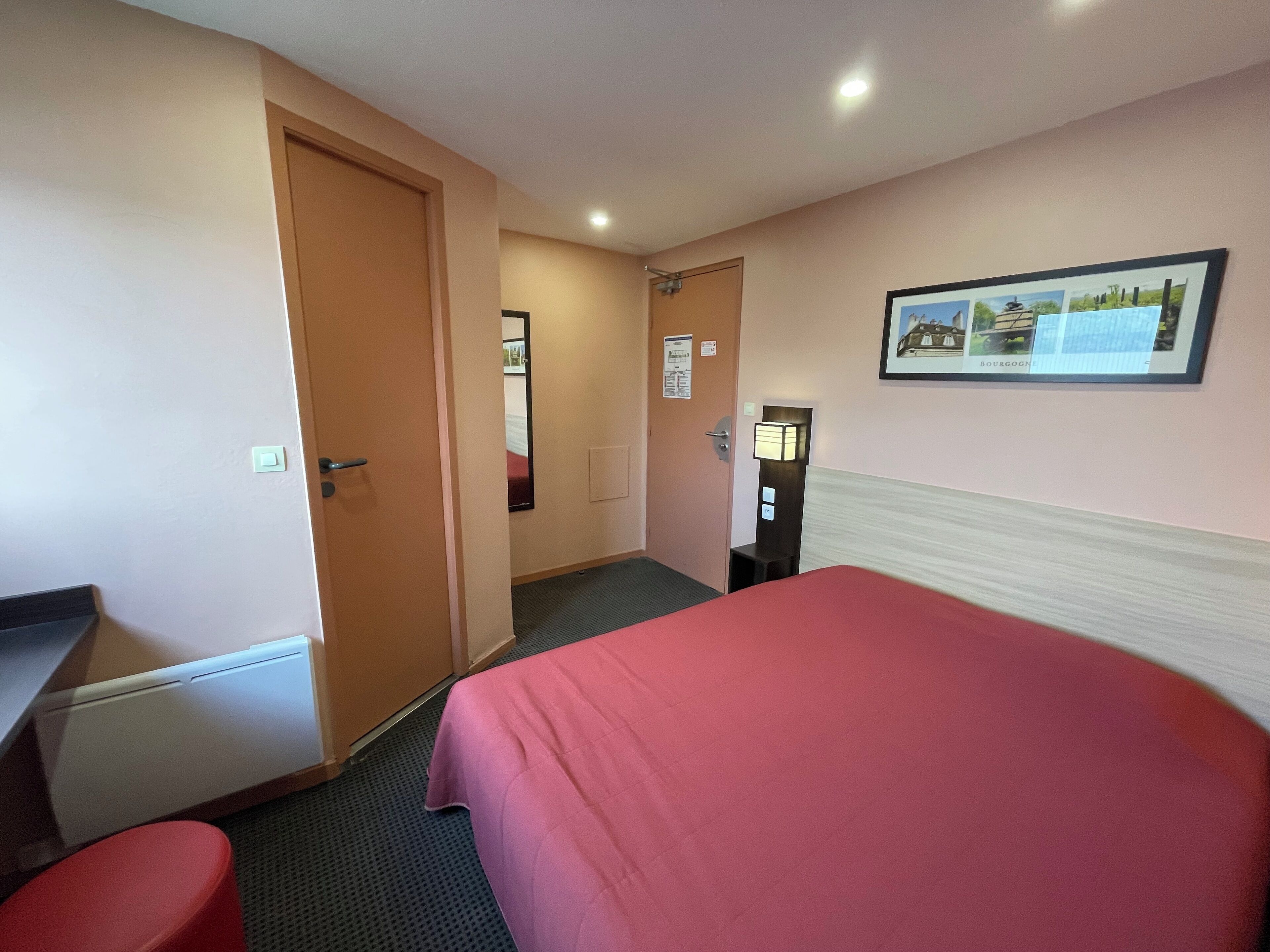 A hotel room with a bed, a desk, a chair, a nightstand, a wall-mounted lamp, and a framed picture.