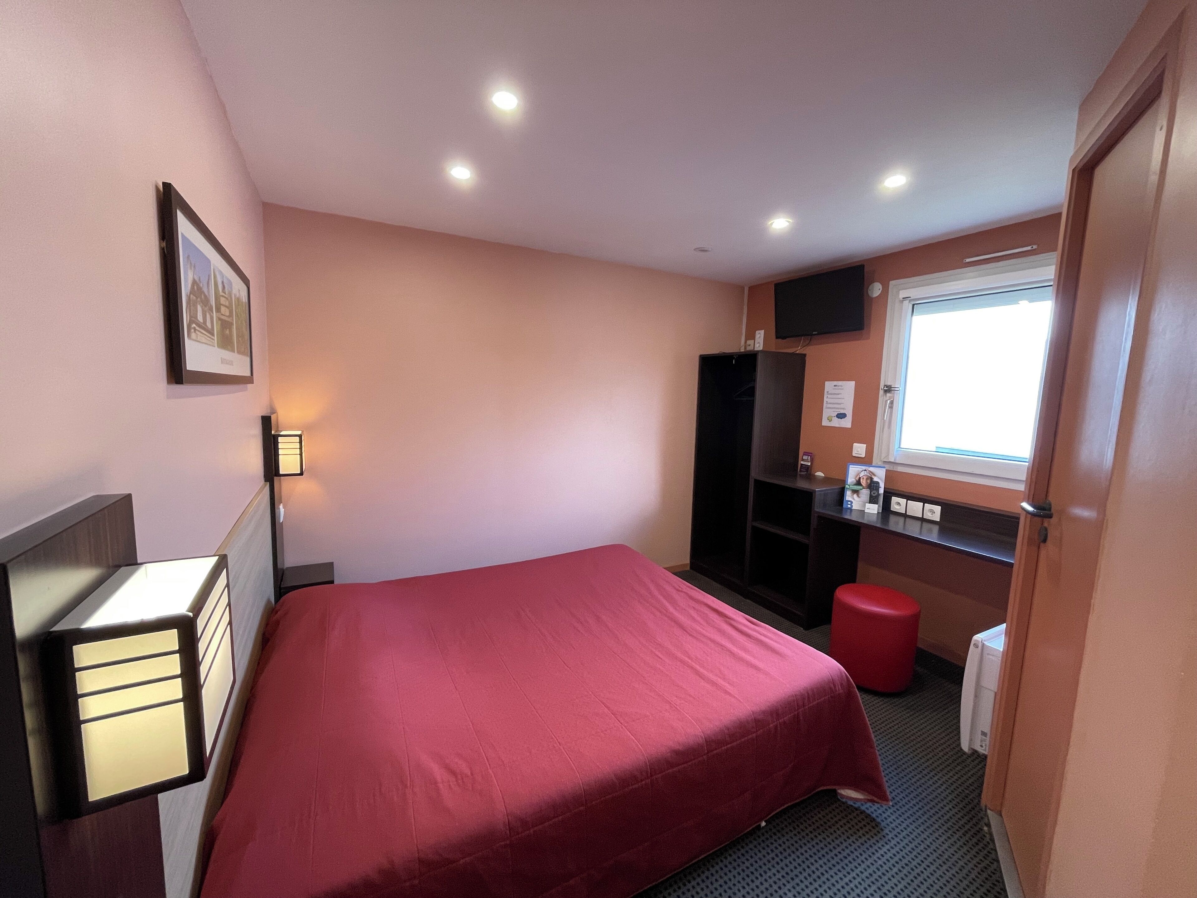 A small, neatly arranged bedroom with a single bed, a wardrobe, a desk with a chair, a window, and a red stool.