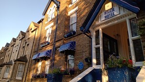 Un edificio de ladrillo tradicional con un letrero que indica el Hotel River. Cuenta con múltiples ventanas, algunas con toldos, y macetas con flores en los alféizares.