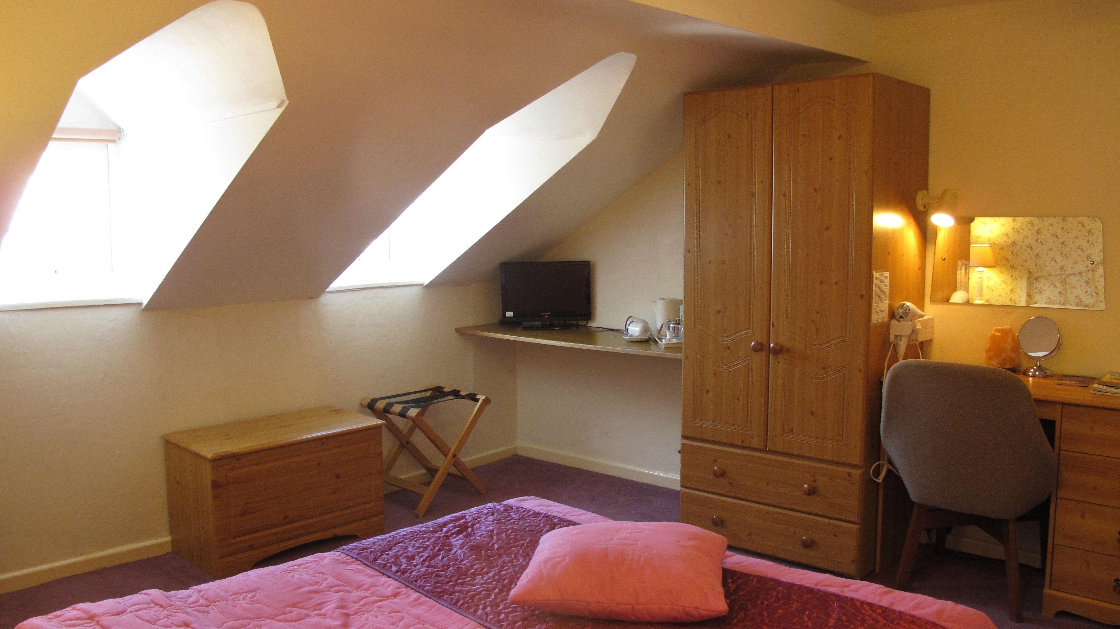 A bedroom with a sloped ceiling, a bed with a pink blanket, a wooden dresser, a desk with a computer, and a chair.