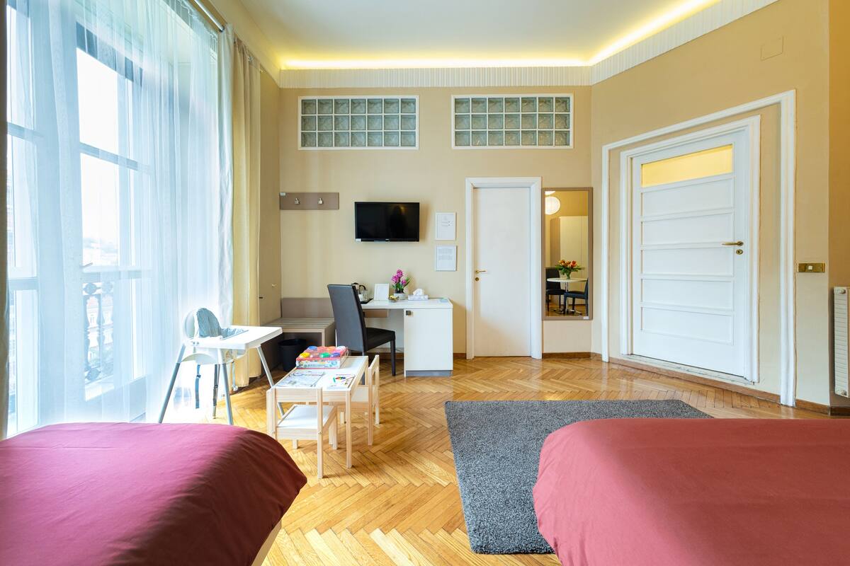 Quarto de hotel com cama, escrivaninha, cadeira, televisão e vista da cidade através de grandes janelas.