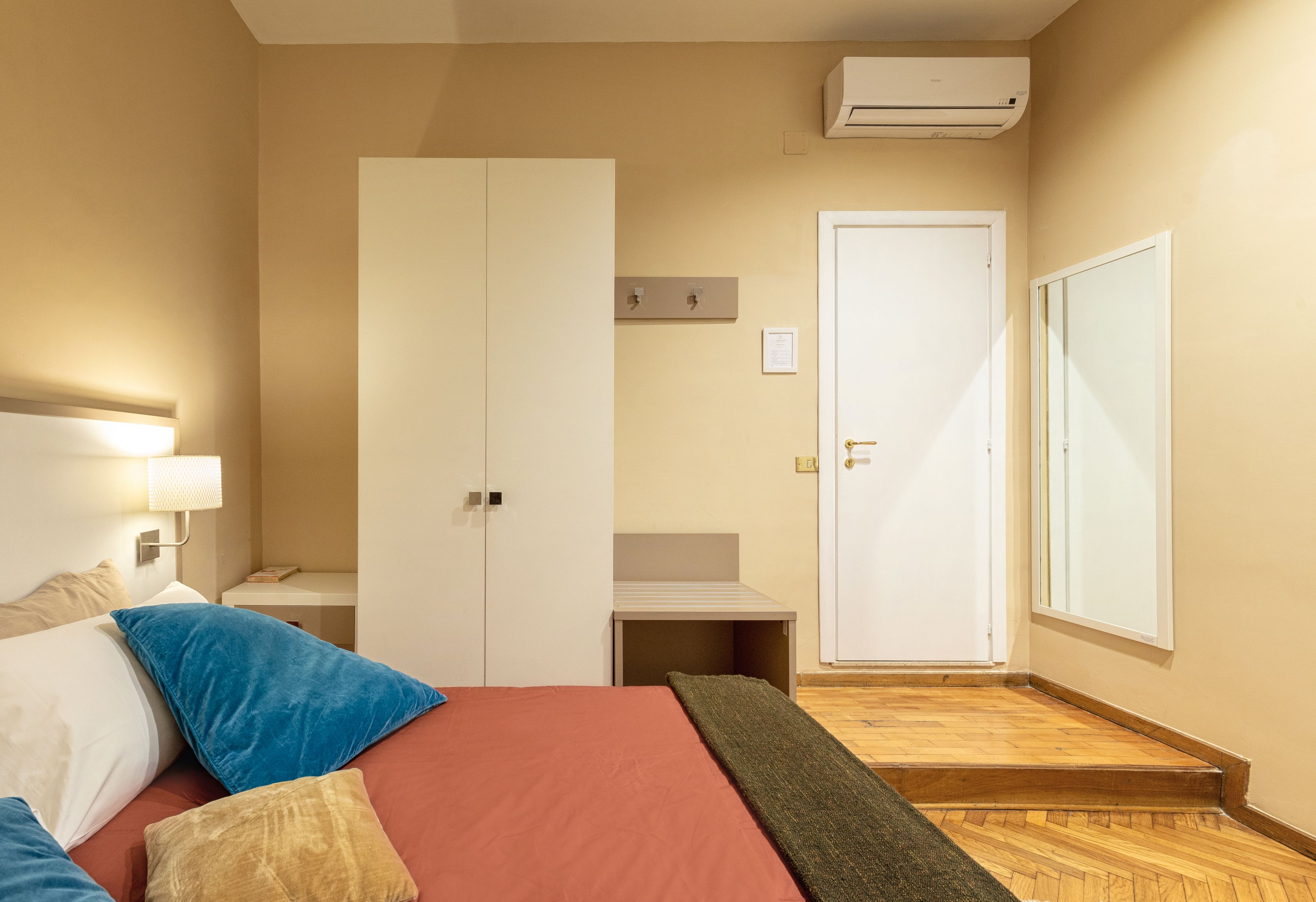 A hotel room with a bed, a bedside table, a wardrobe, a door, and a small desk.