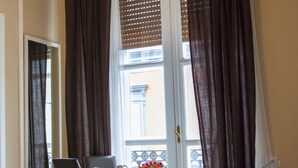 Habitación de hotel con una cama, un escritorio con una silla, una mesita con un jarrón de flores y una ventana con cortinas.