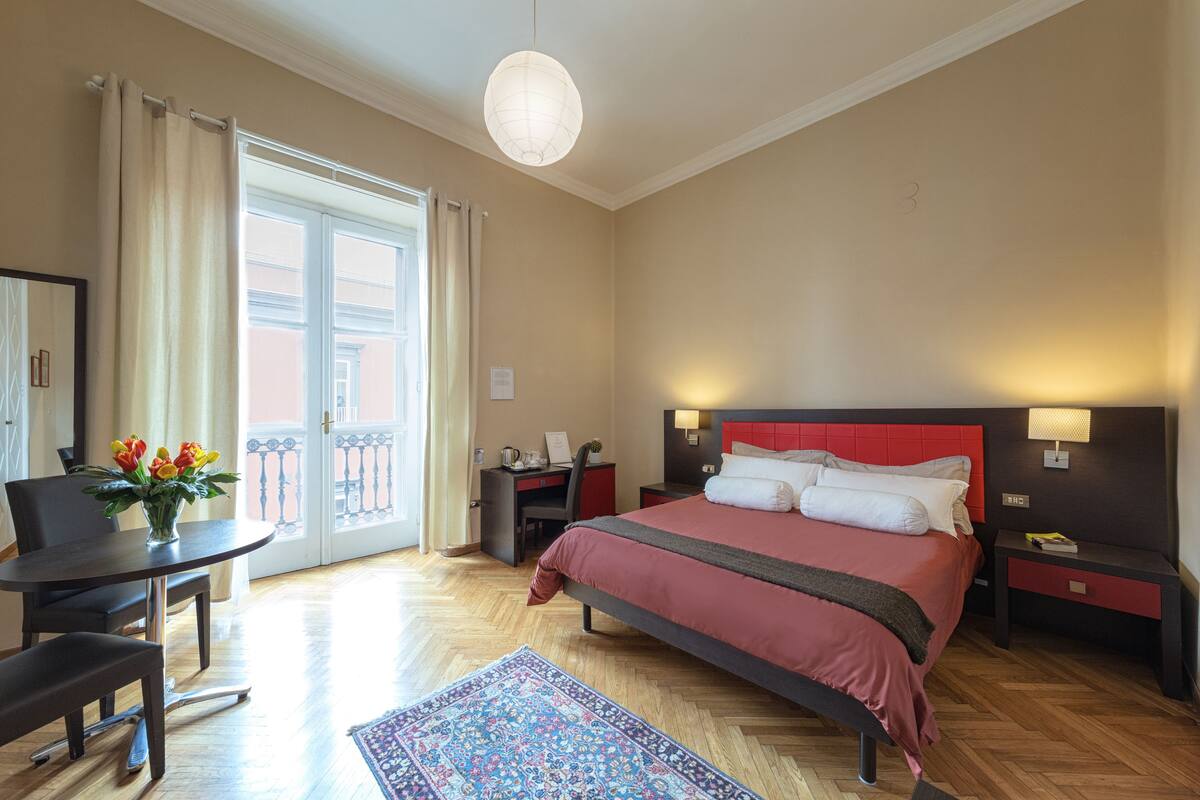 Quarto de hotel com cama, mesas de cabeceira, escrivaninha, cadeira, mesa de jantar e janela com cortinas.