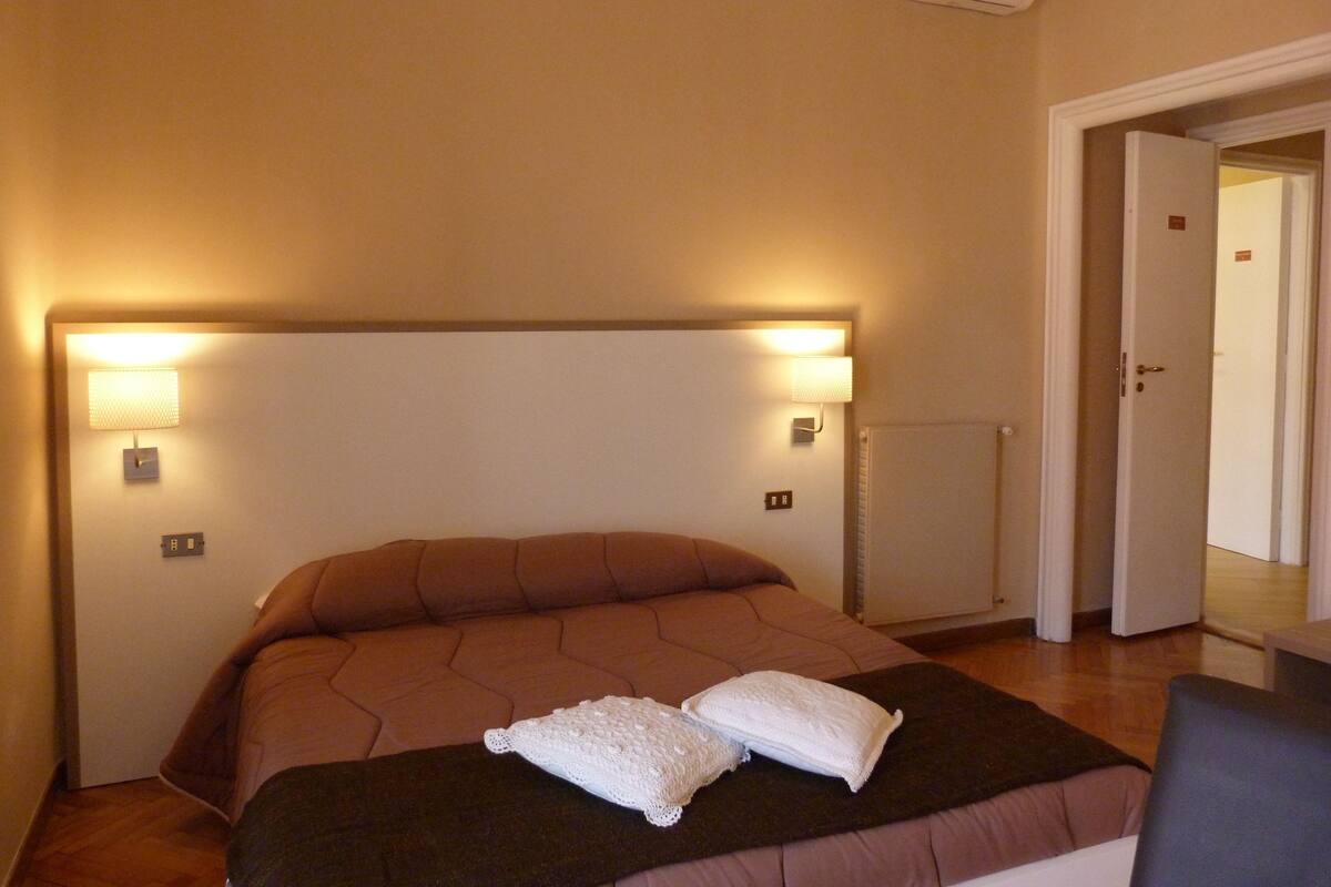 Quarto de hotel com uma cama grande, duas luminárias de cabeceira, um termostato e uma porta que dá para outro cômodo.
