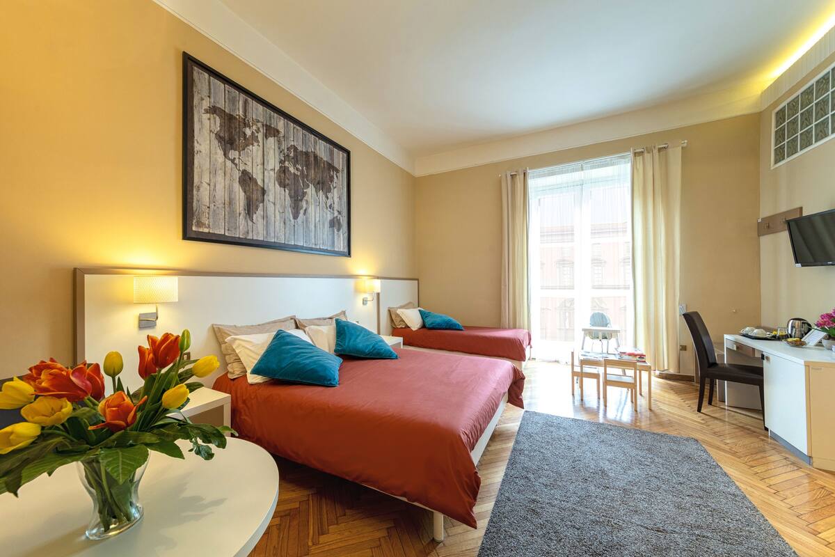 Quarto de hotel com duas camas, uma escrivaninha, uma cadeira, televisão e janela com cortinas.