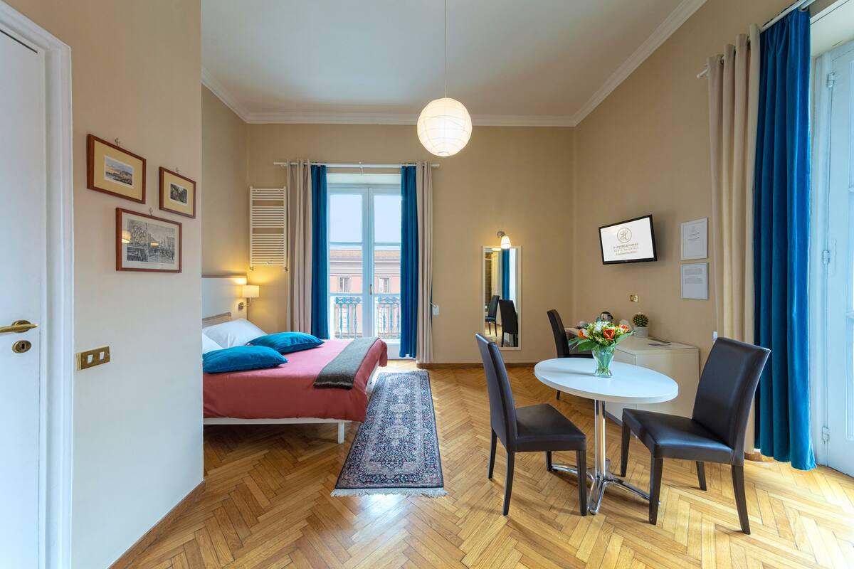 Quarto de hotel com cama, secretária e cadeiras, janela com cortinas e televisão fixada na parede.