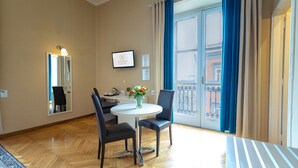 Habitación de hotel con zona de comedor que incluye una mesa redonda, dos sillas, un florero con flores y un amplio ventanal con cortinas.
