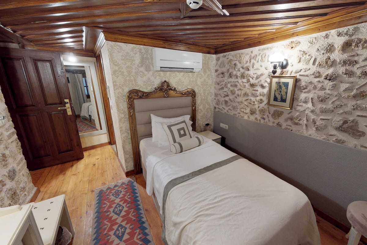 Een slaapkamer met een houten plafond, een stenen muur, een bed met hoofdeinde, een nachtkastje en een klein krukje.