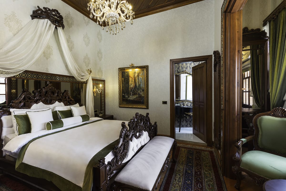 Een groot, versierd bed met een hemelbed, een kroonluchter en een schilderij aan de muur.