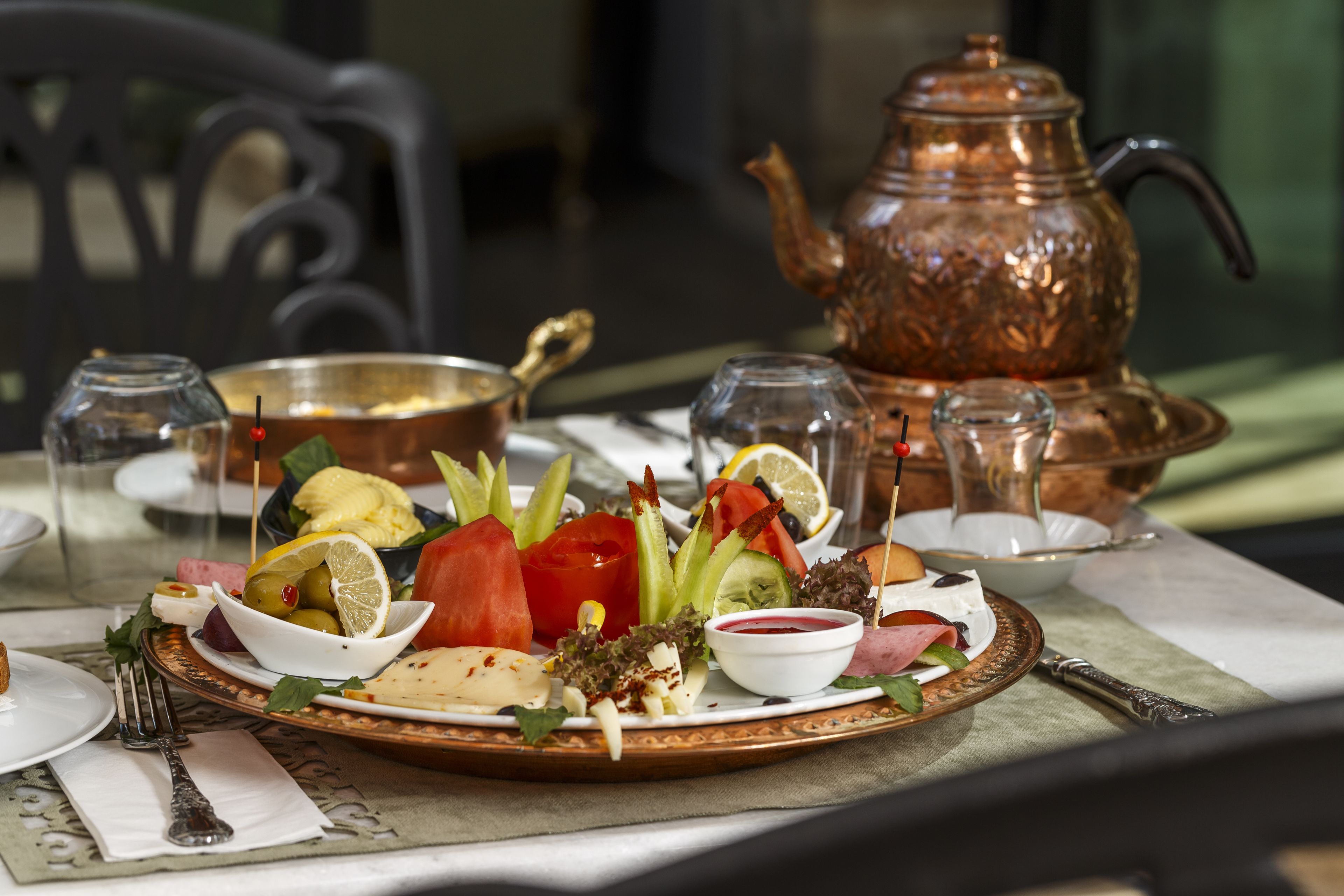 A table set with a variety of dishes, including a salad, a bowl of olives, and a plate with a slice of tomato. There is a copper teapot and a glass pitcher on the table.