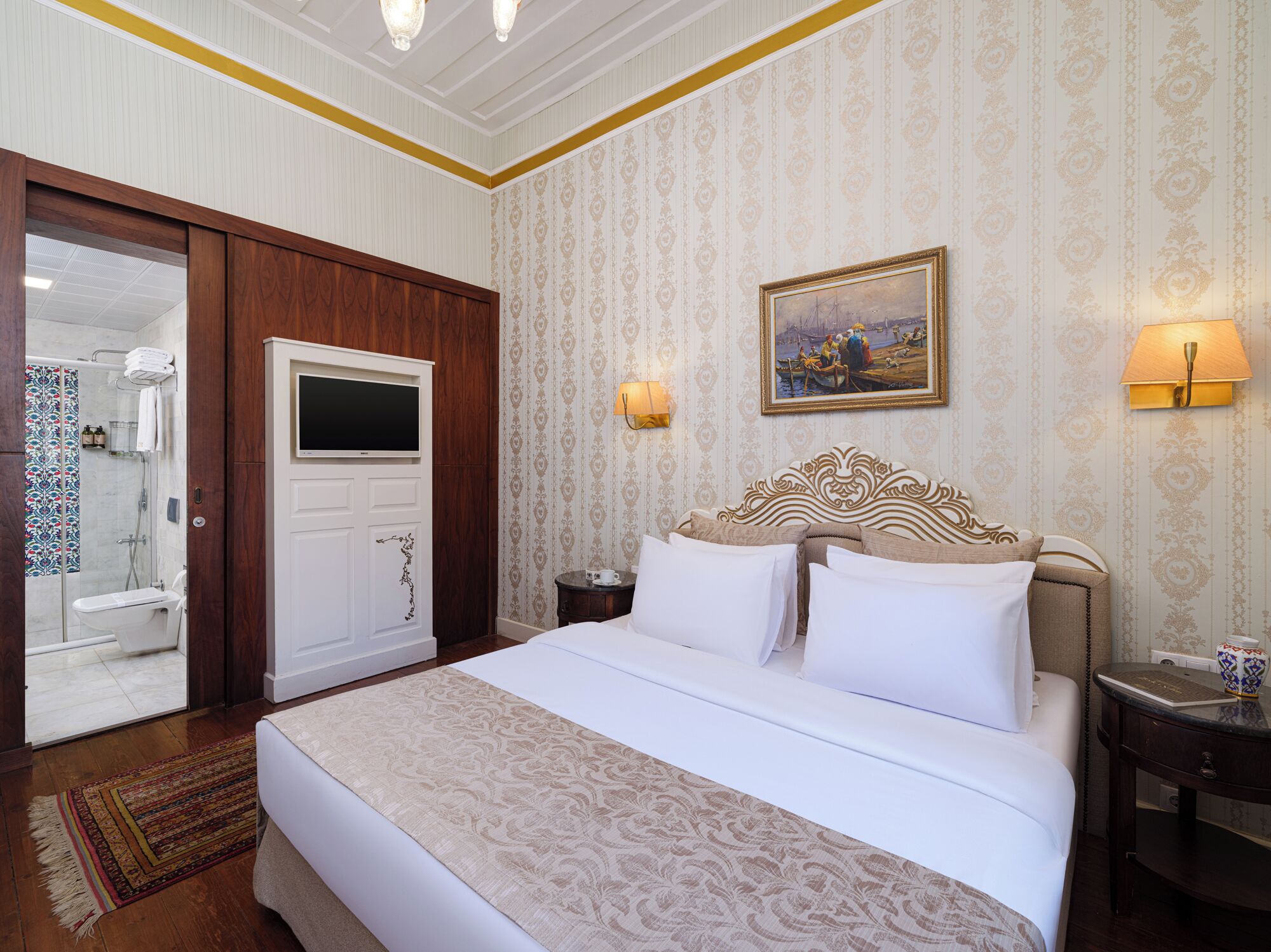 Quarto de hotel com uma cama grande, televisão, pintura na parede e banheiro visível por uma porta aberta.