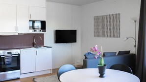 Une cuisine moderne équipée d’un four à micro-ondes, d’un four et d’un évier. On y trouve une table à manger avec des chaises et un pot d’orchidée. Un téléviseur à écran plat est fixé au mur, et un canapé est visible au fond de la pièce.