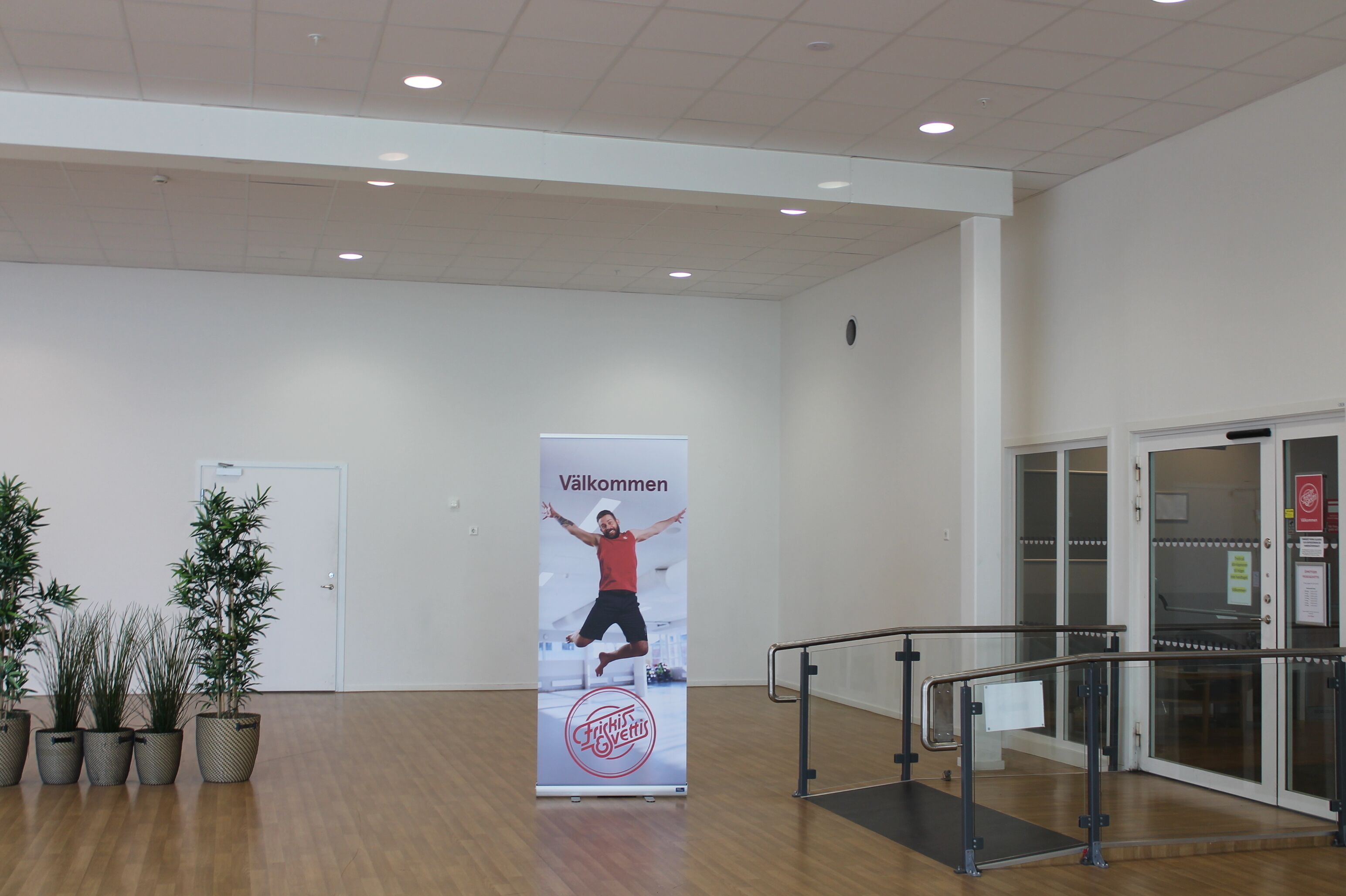 A spacious indoor area with a wooden floor, a large vertical banner with text, potted plants, and a glass door.