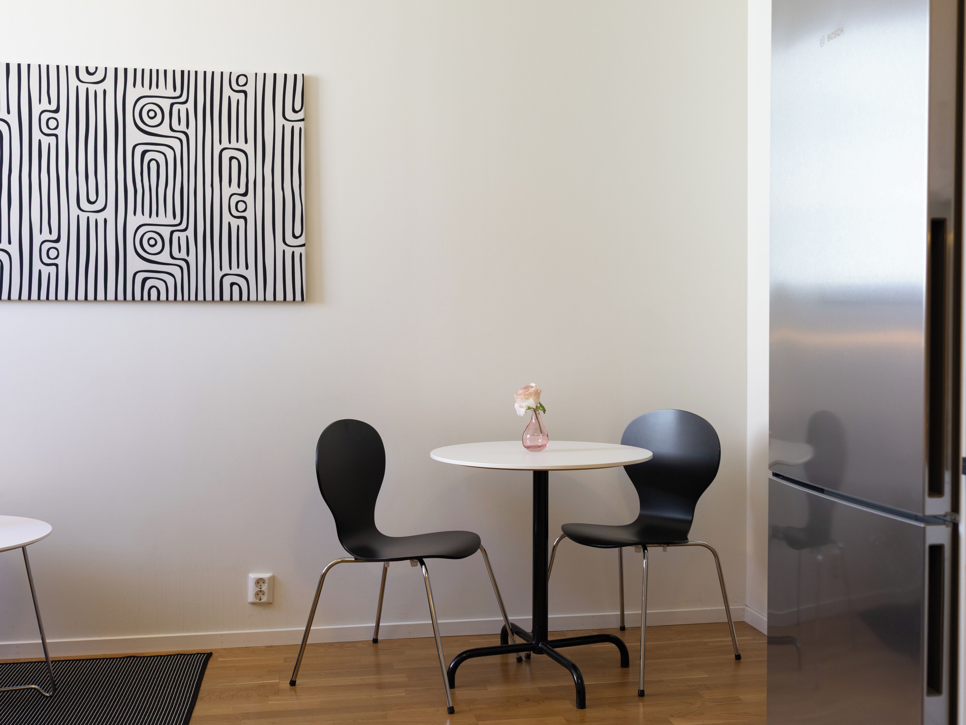 A modern room with a round table, two black chairs, a vase with a single flower, and a wall-mounted abstract art piece.