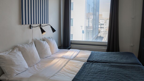 Une chambre d’hôtel avec un lit, une fenêtre avec des rideaux, une lampe fixée au mur et une œuvre d’art à rayures.