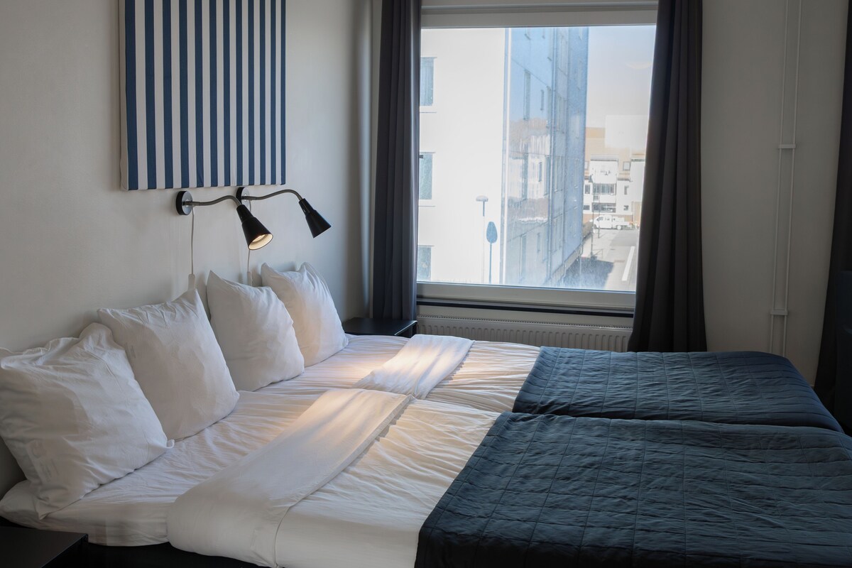 Ein Hotelzimmer mit Bett, einem Fenster mit Vorhängen, einer Wandlampe und einem gestreiften Kunstwerk.