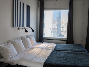 Una habitación de hotel con una cama, una ventana con cortinas, una lámpara de pared y una obra de arte a rayas.
