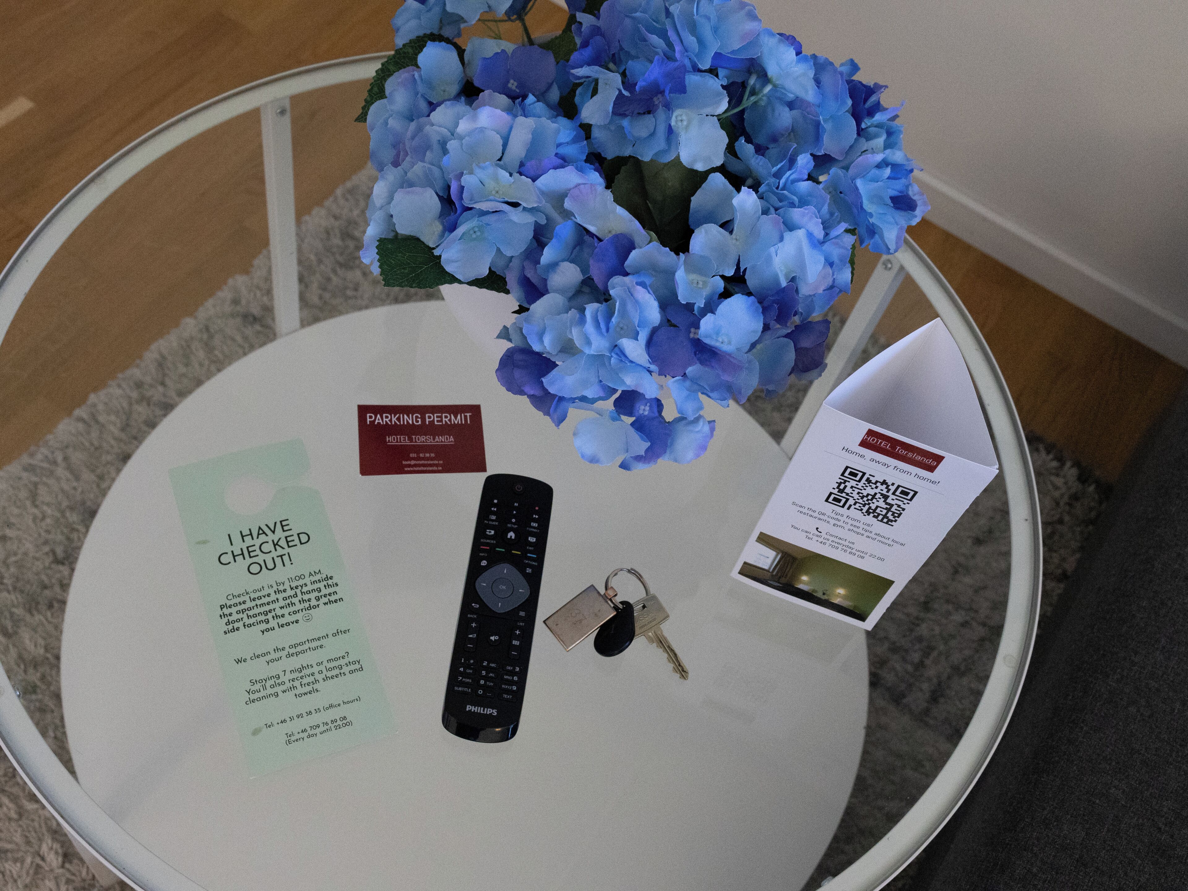 Ein Glastisch mit Fernbedienung, Schlüsseln, Parkausweis und einem QR-Code auf einer Hotelbroschüre.