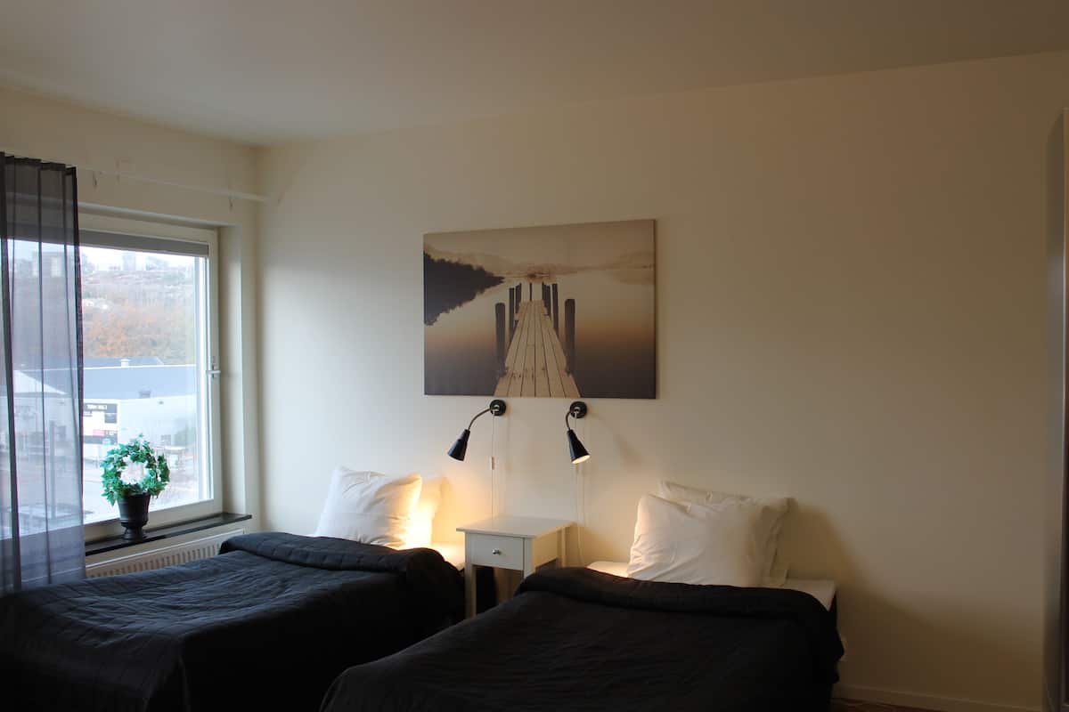 Ein Hotelzimmer mit zwei Betten, einem Nachttisch, einer Lampe und einem Fenster mit Blick auf Gebäude.