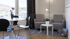 Une pièce moderne avec une table à manger ronde, deux fauteuils gris, une petite table blanche, un lampadaire et une fenêtre avec des rideaux.