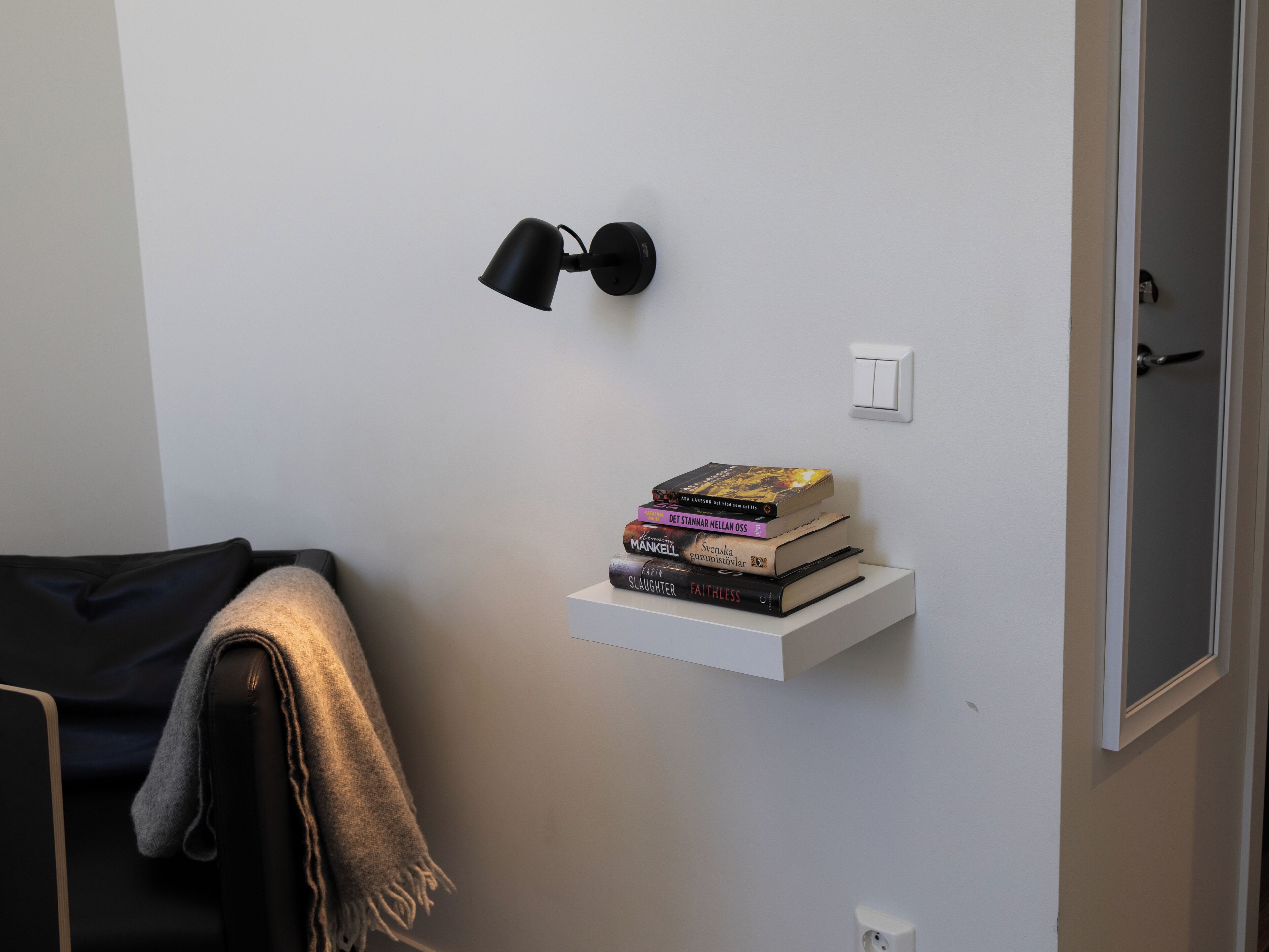 Uma parede branca com um candeeiro de parede preto, uma estante branca com livros, uma porta e um casaco pendurado numa cadeira.