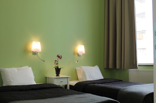 Una habitación de hotel con dos camas, paredes verdes, una mesita de noche con lámpara y una ventana con cortinas.
