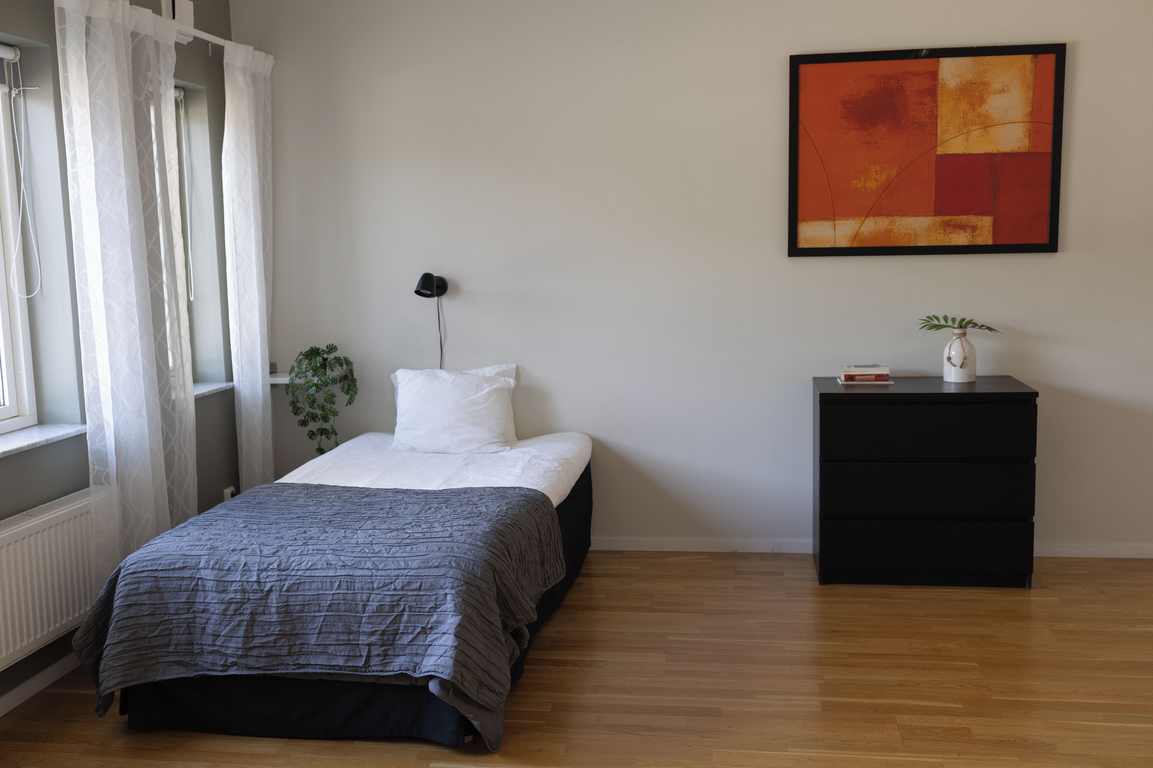 Een slaapkamer met een bed, nachtkastje, lamp, plant en een schilderij aan de muur.