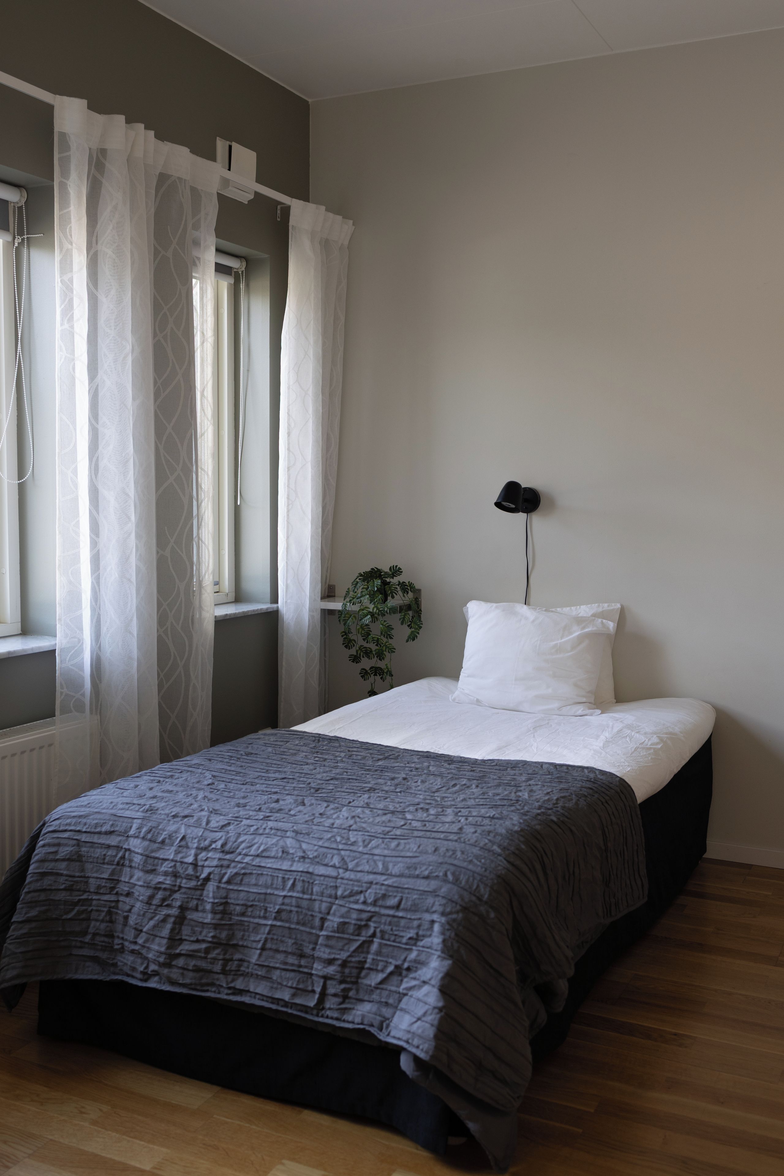 A bedroom with a bed, a window with sheer curtains, a bedside lamp, and a potted plant.
