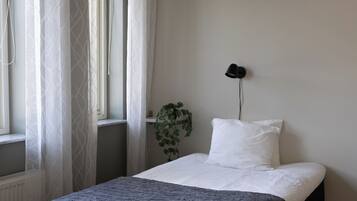 A bedroom with a bed, a window with sheer curtains, a bedside lamp, and a potted plant.