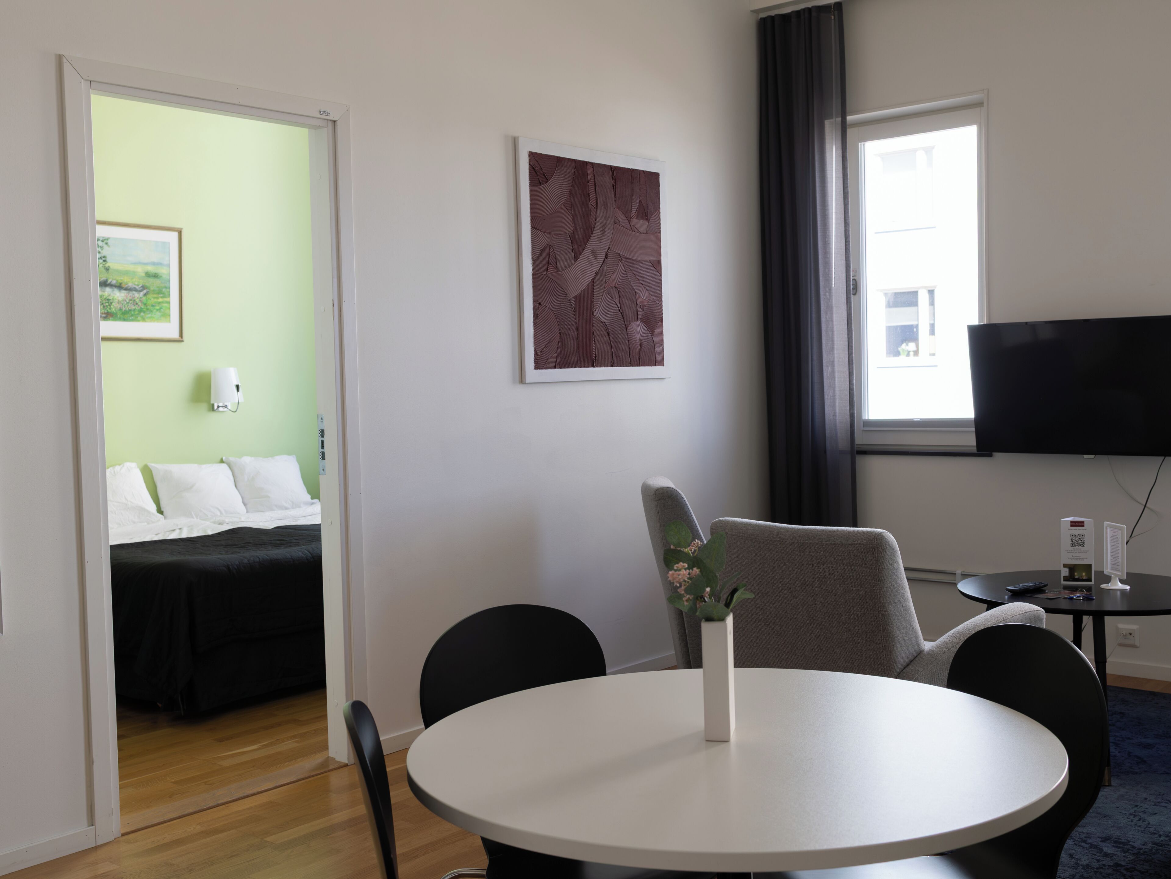 A hotel room with a bed, a round dining table, a chair, a small table with a vase, a flat-screen TV, and a window with curtains.