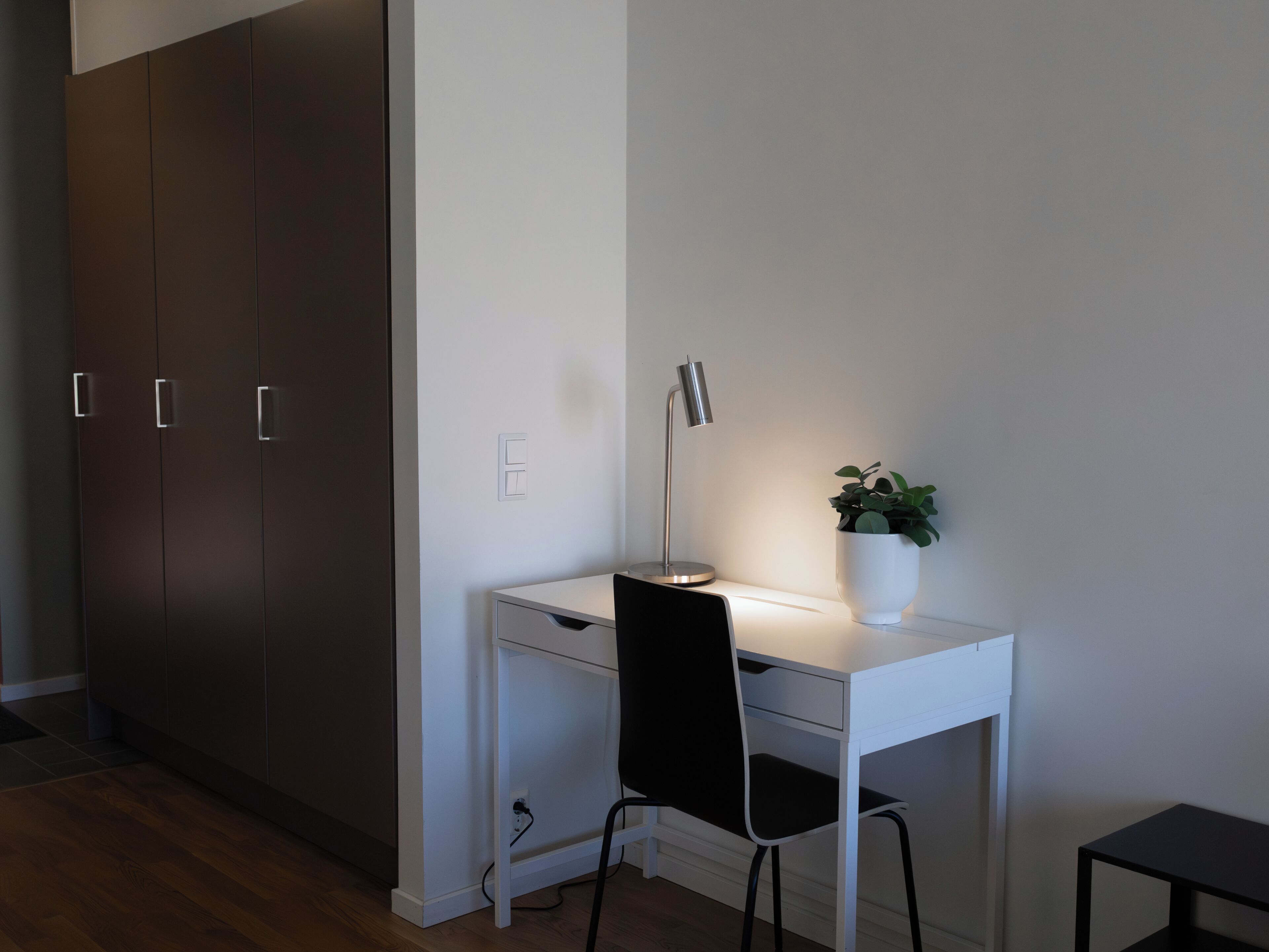 Ett vitt skrivbord med en svart stol, en skrivbordslampa och en krukväxt.