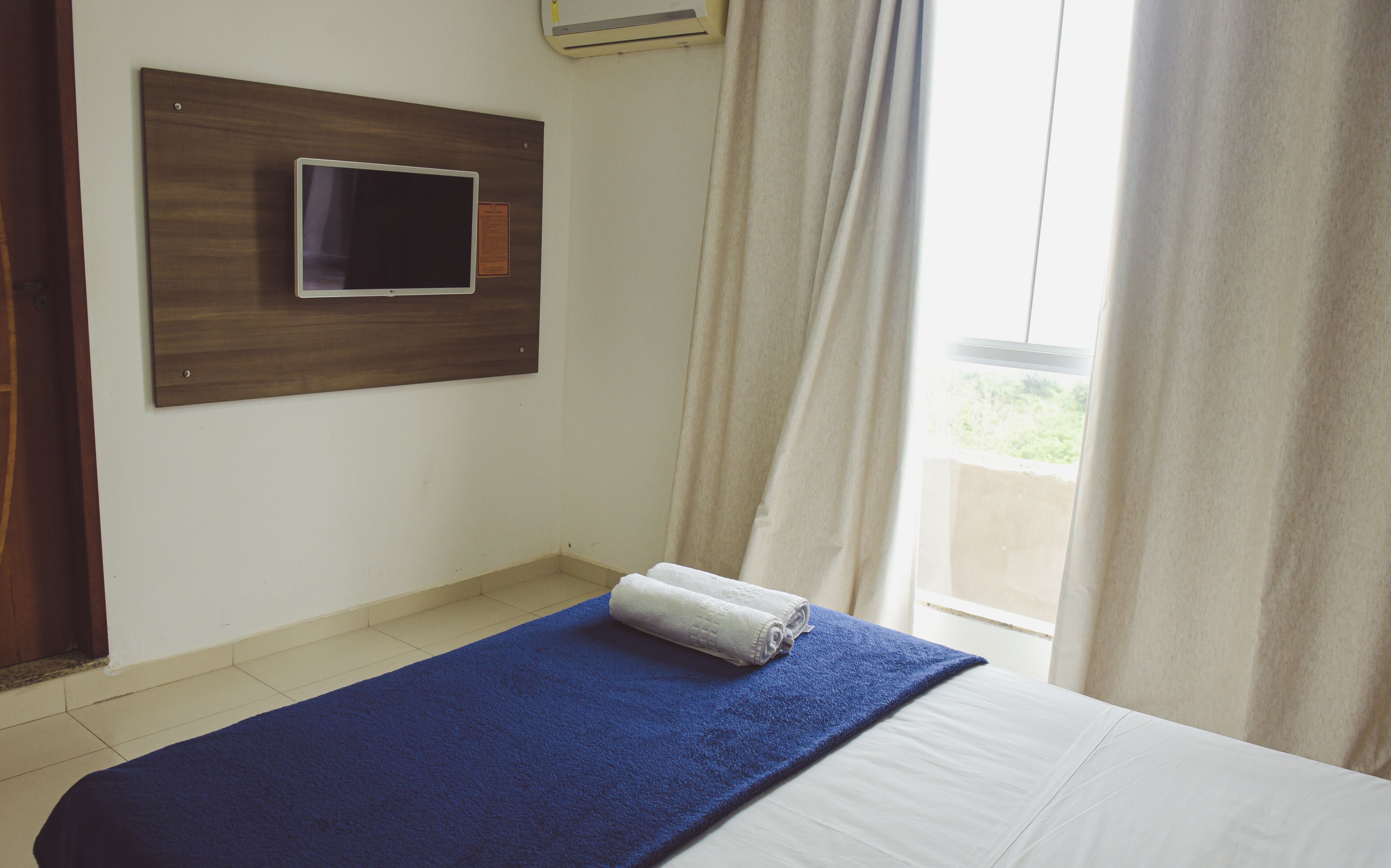 A hotel room with a bed, a mounted television, and a view of the outdoors through a window.