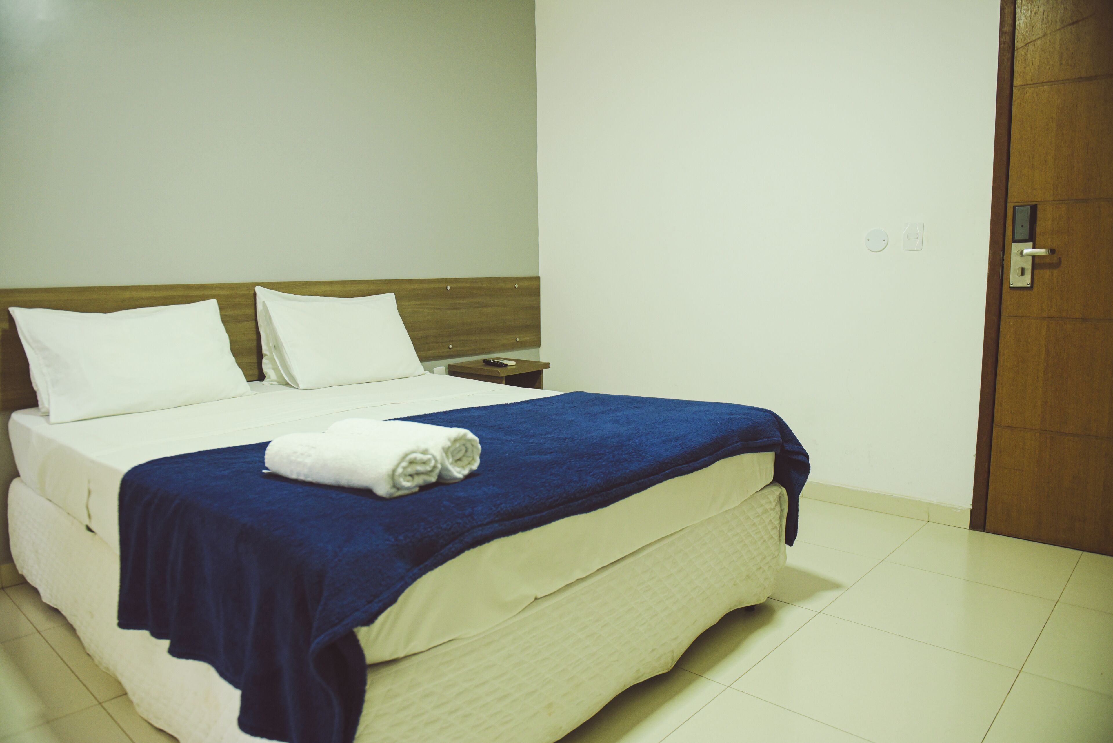 Una cama bien hecha con un edredón azul y dos almohadas blancas, un cabecero de madera, una mesita de noche con un control remoto y una puerta con manija.
