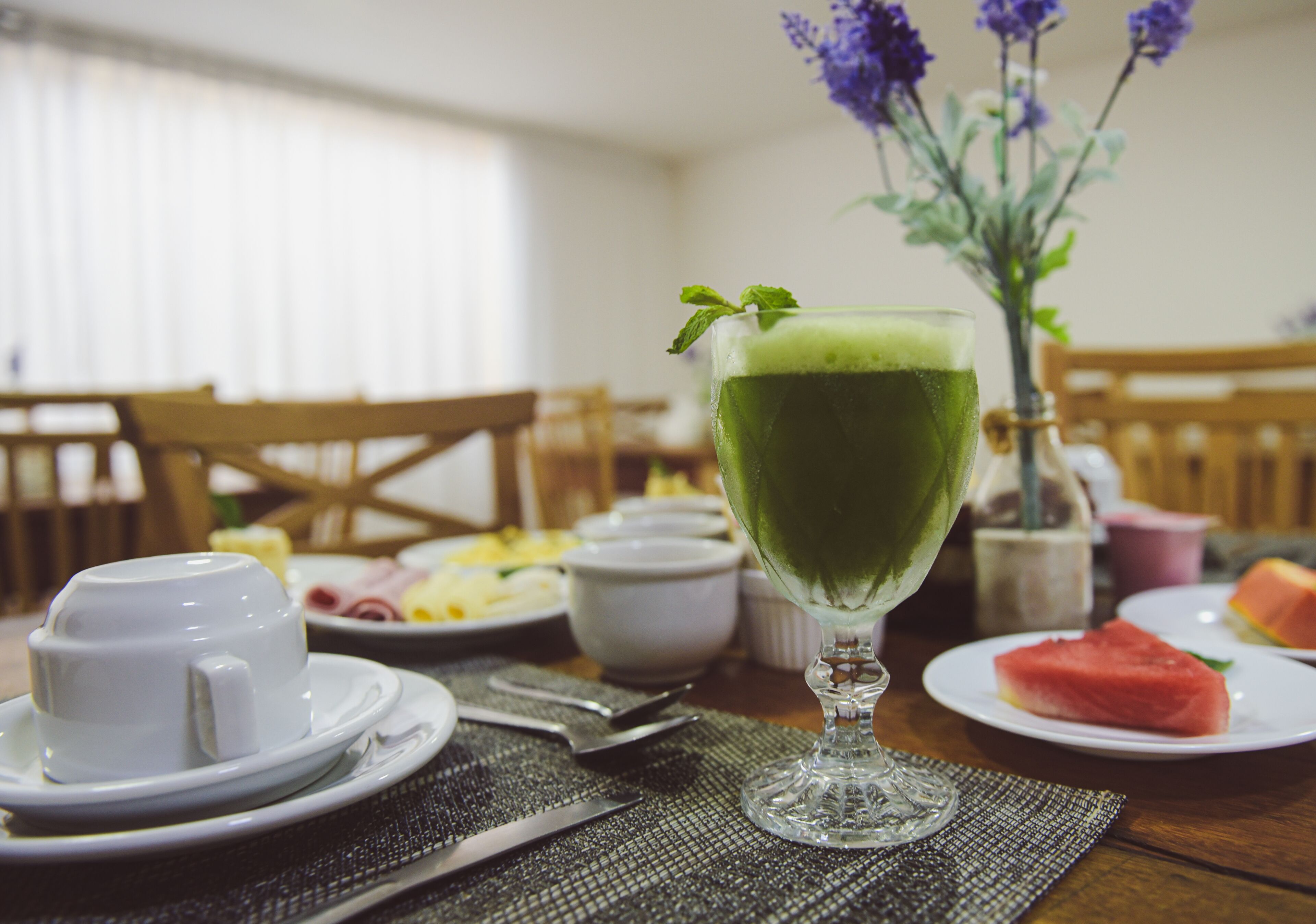 A table set with a green beverage in a glass, white cups, a plate with watermelon, and a vase with purple flowers.