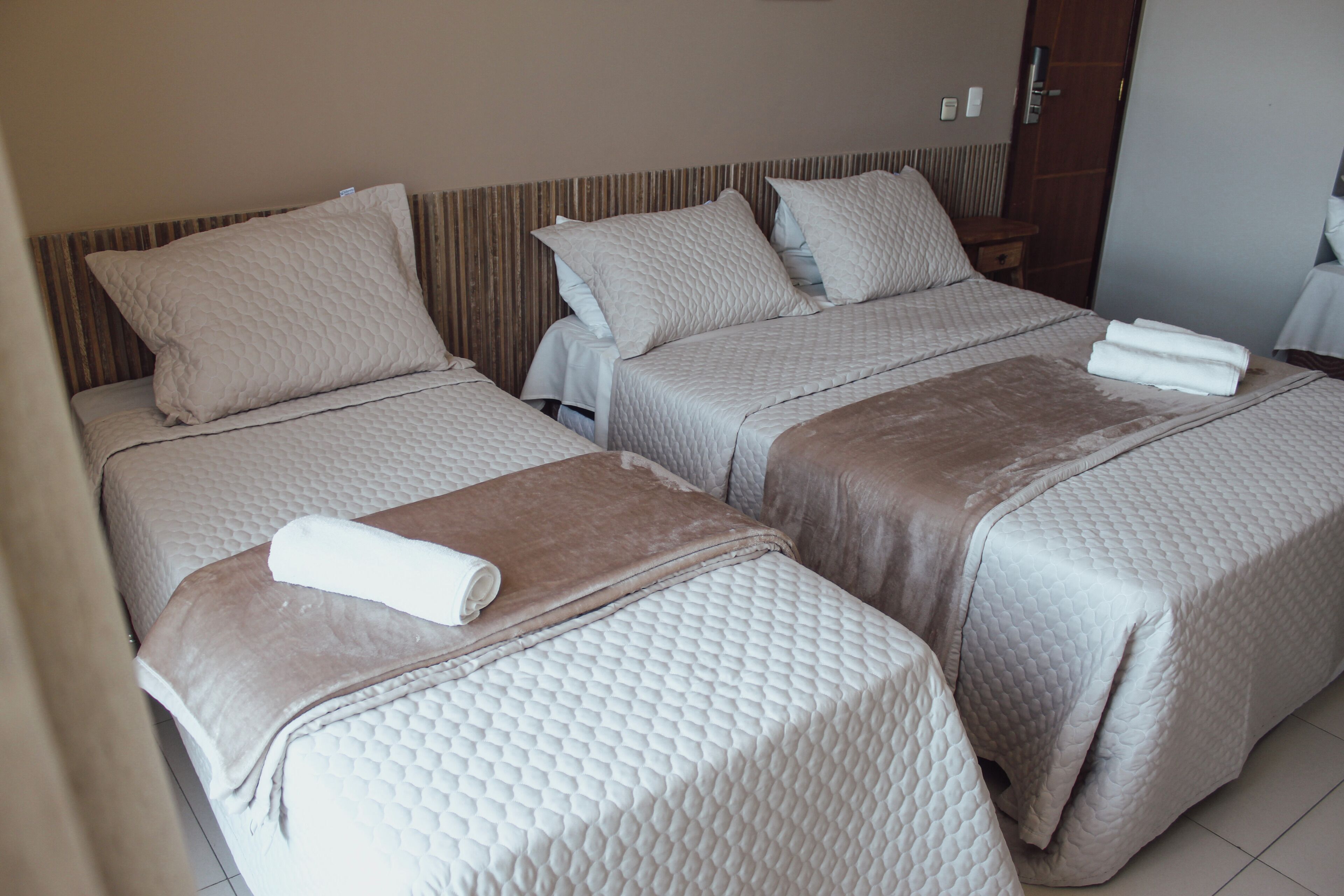 Habitación de hotel con dos camas, cada una con colchón acolchado y almohadas, y una manta marrón sobre una de ellas.