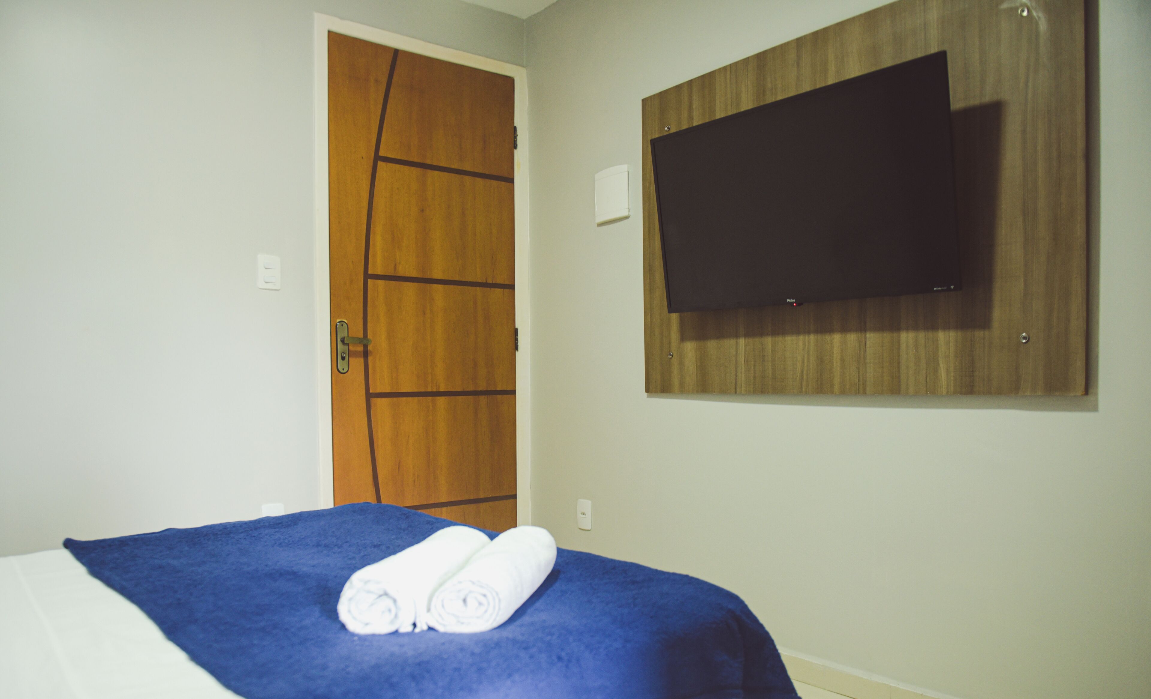A hotel room with a bed, a wooden door, a flat-screen TV mounted on the wall, and a towel on the bed.