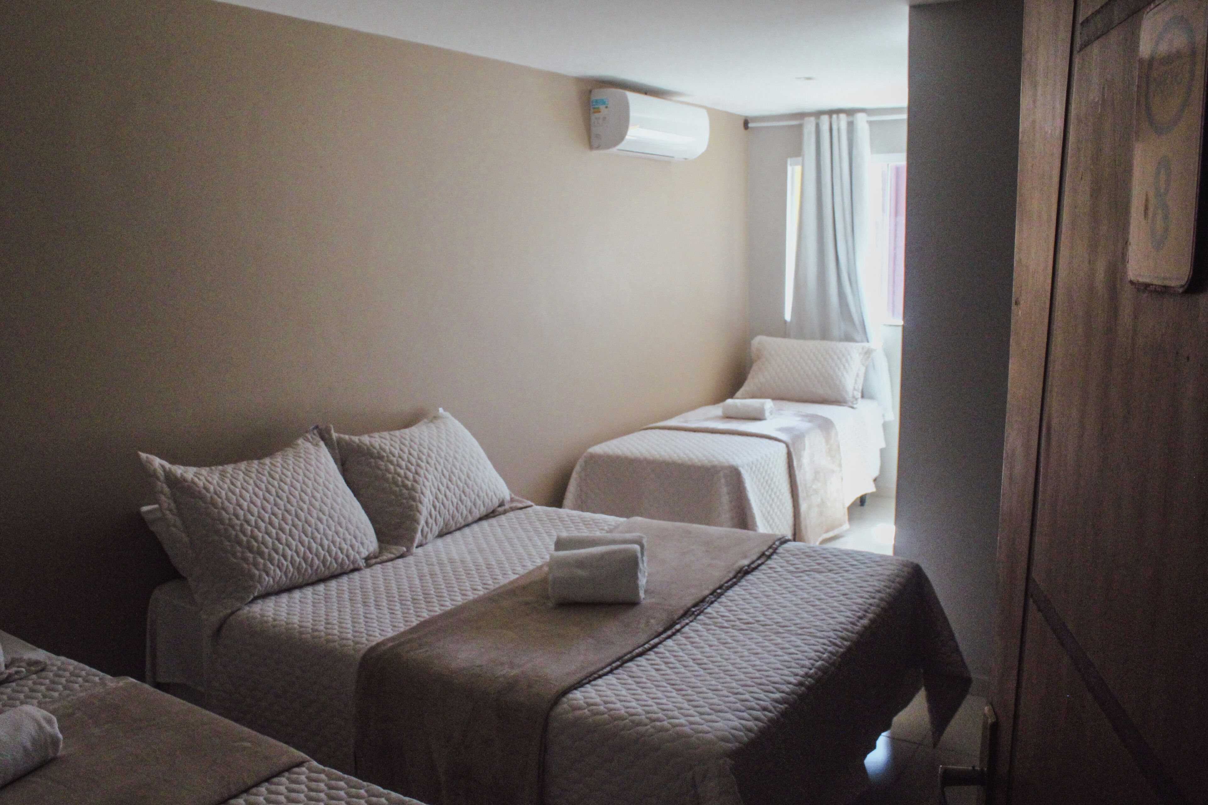 A hotel room with three beds, each with a quilted bedspread and pillows. There is an air conditioning unit on the wall and a wooden wardrobe.