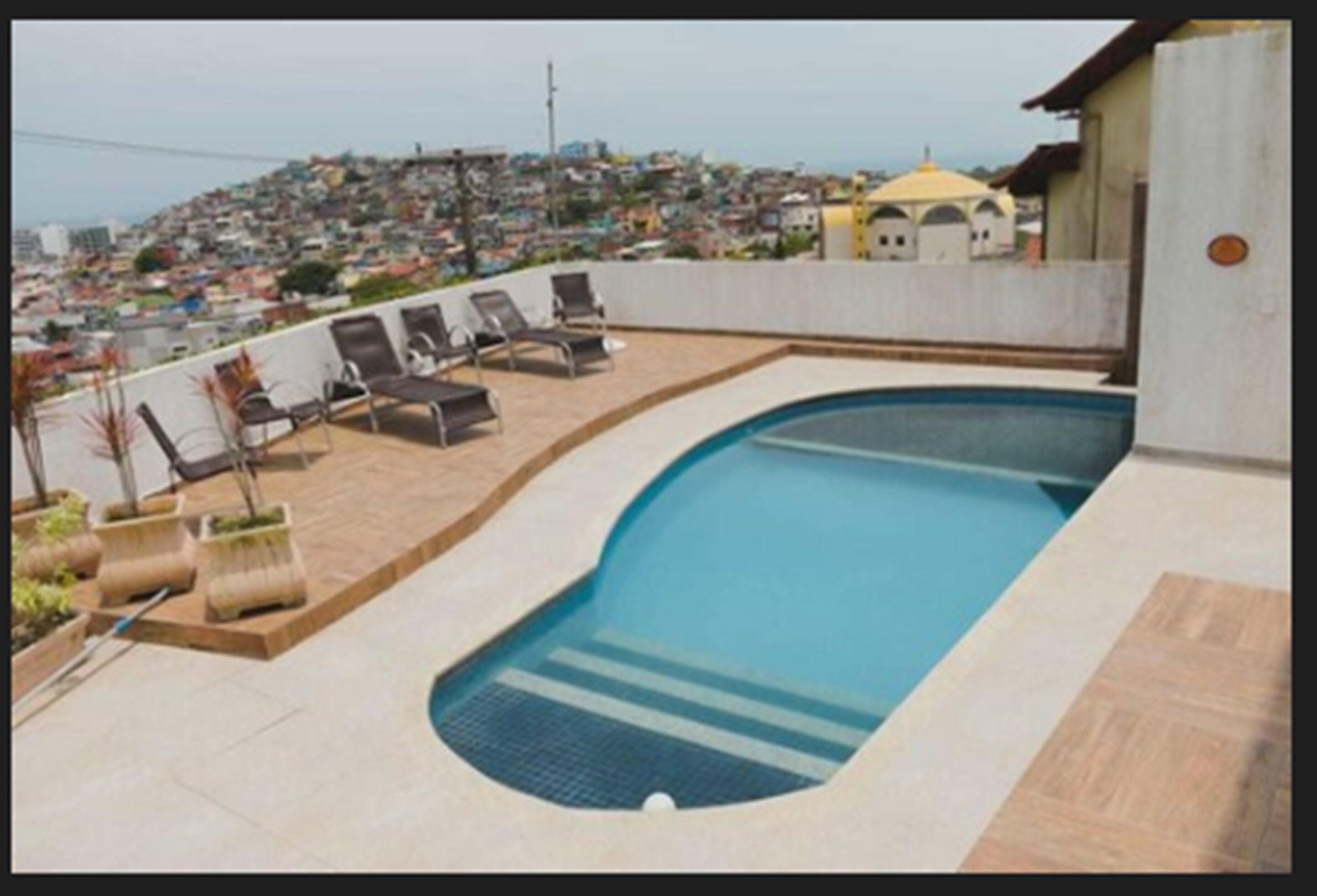 A rooftop swimming pool with lounge chairs and a view of a densely populated hillside.
