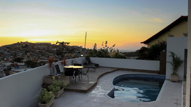 A rooftop terrace with a swimming pool, outdoor furniture, and a view of a densely packed cityscape during sunset. - Pousada Xerxes (Arraial do Cabo)