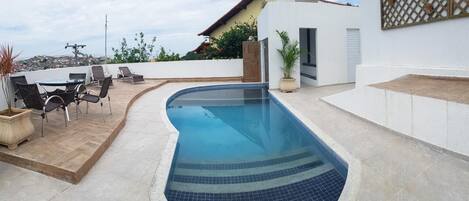 A swimming pool with a tiled edge, surrounded by a patio area with chairs and a table.