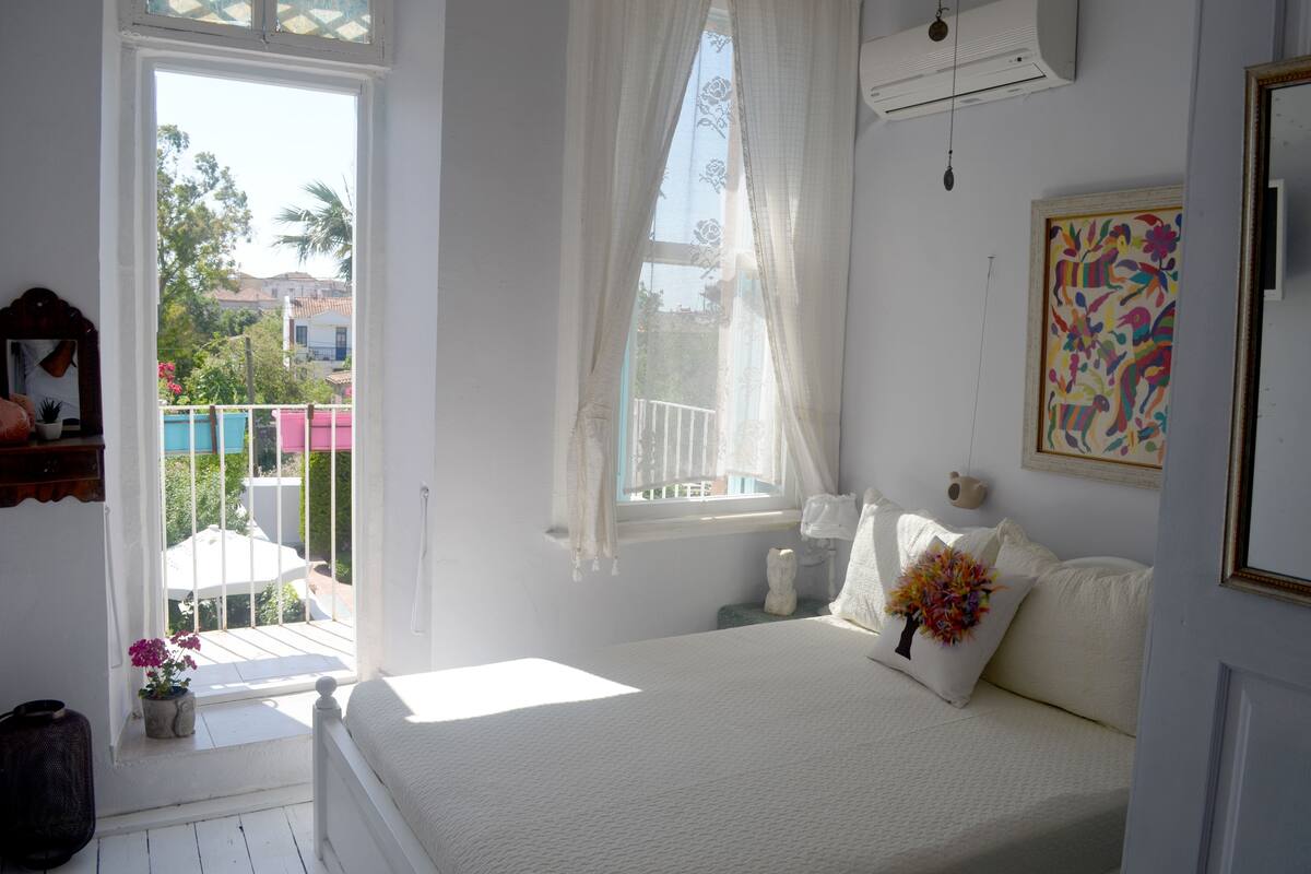 Un dormitorio con cama, ventana con cortinas traslúcidas, un balcón pequeño y un cuadro colorido en la pared.