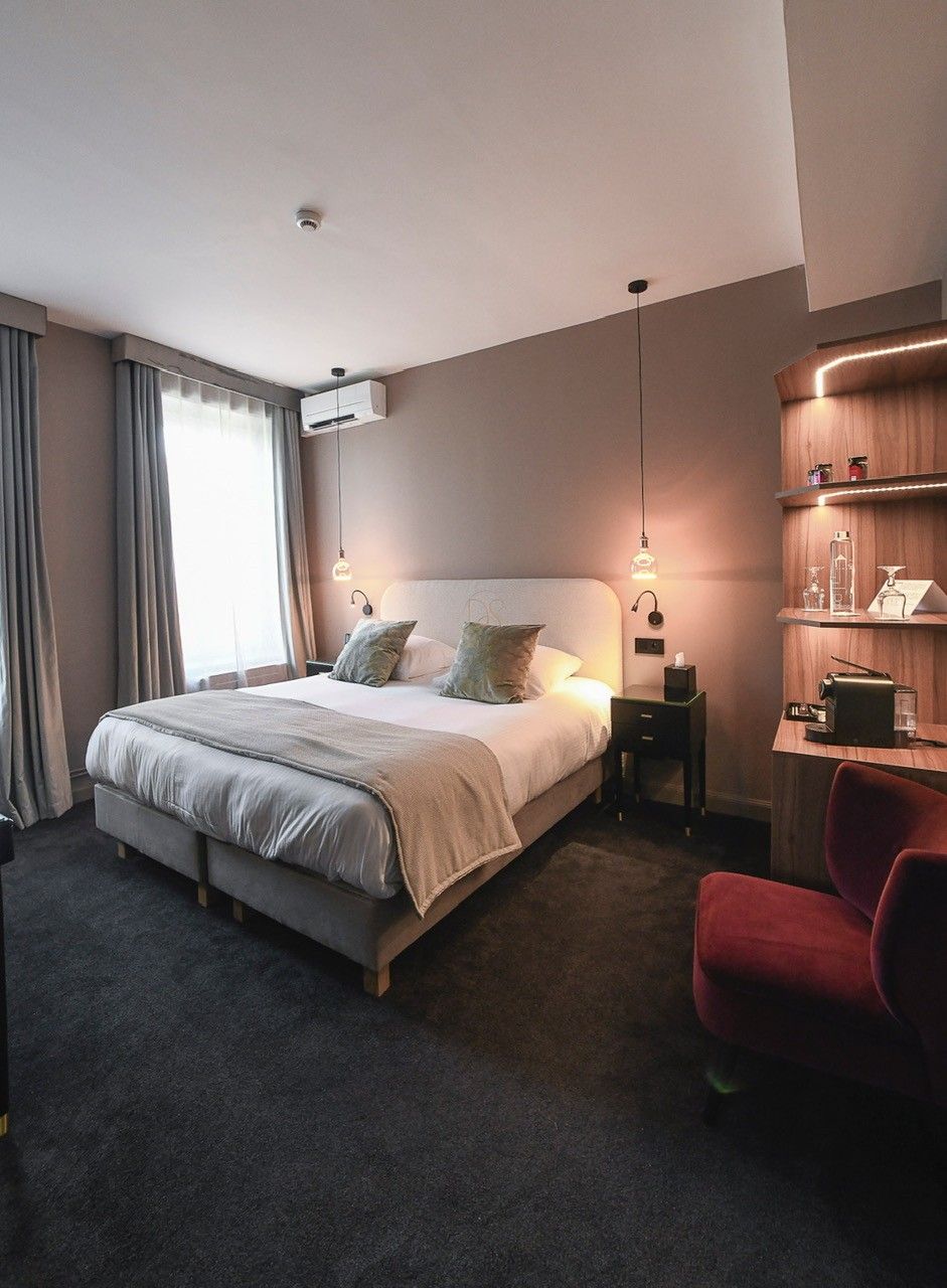 Una habitación de hotel moderna con una cama grande, mesitas de noche, un sillón rojo y un estante de madera con objetos.